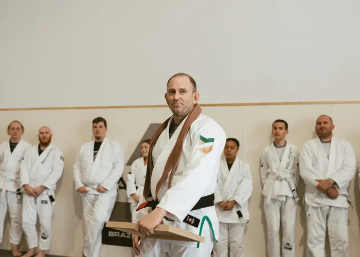 A Balance BJJ coach in a brown belt holding a grading board with members lined up behind